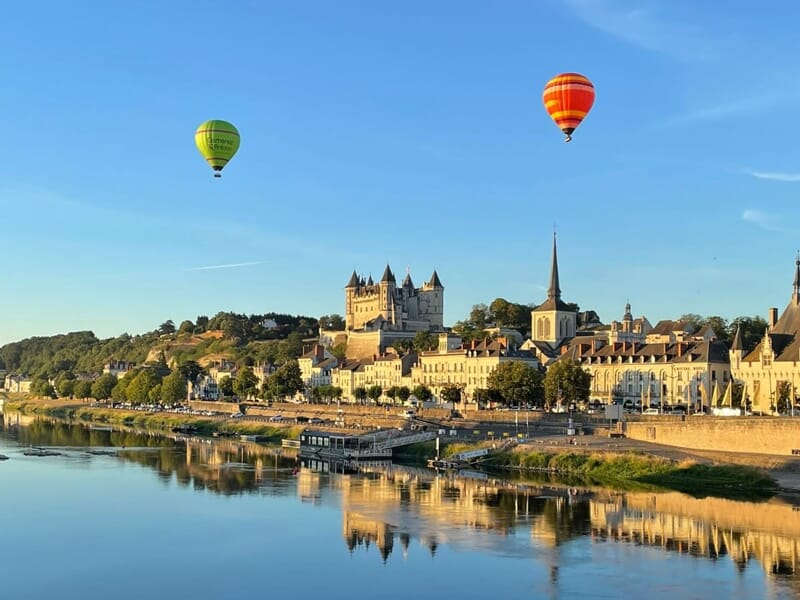 Deux montgolfières colorées au-dessus d'une ville historique avec un château et une église près d'une rivière.
