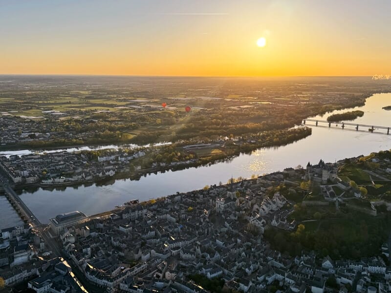 Vue panoramique au coucher du soleil d'une ville avec une rivière, un pont et deux montgolfières dans le ciel.