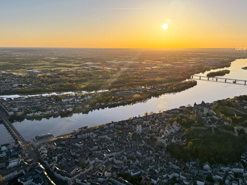 Vue panoramique au coucher du soleil d'une ville avec une rivière, un pont et deux montgolfières dans le ciel.