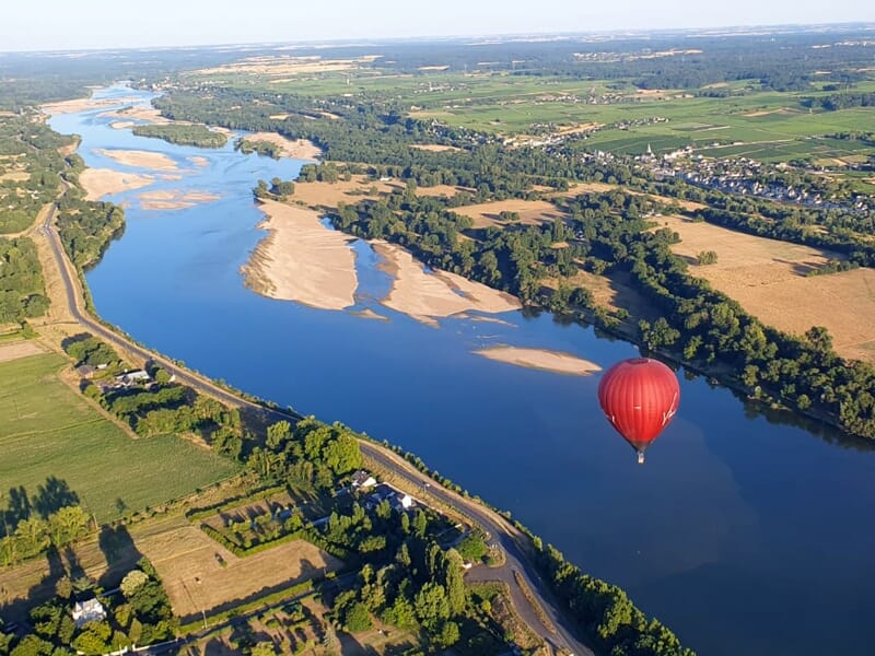 Montgolfière rouge volant au-dessus d'une rivière sinueuse entourée de champs verts et de petites habitations.