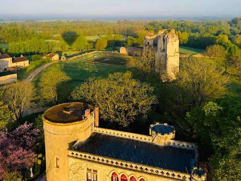 Vue aérienne d'un château ancien entouré d'arbres et de jardins sous un ciel clair au lever du soleil.