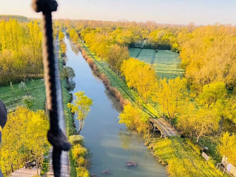 Paysage vu d'une montgolfière avec une rivière bordée d'arbres aux feuilles jaunes et un petit pont en bois.