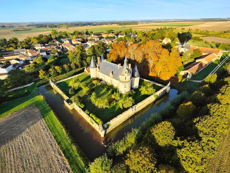 Vue aérienne d'un château entouré d'un jardin et d'un village avec des arbres aux couleurs d'automne.