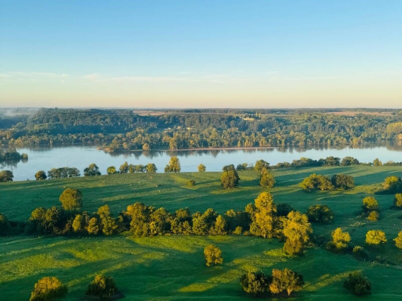 Paysage naturel avec une rivière bordée d'arbres et des champs verts sous un ciel clair au lever du soleil.