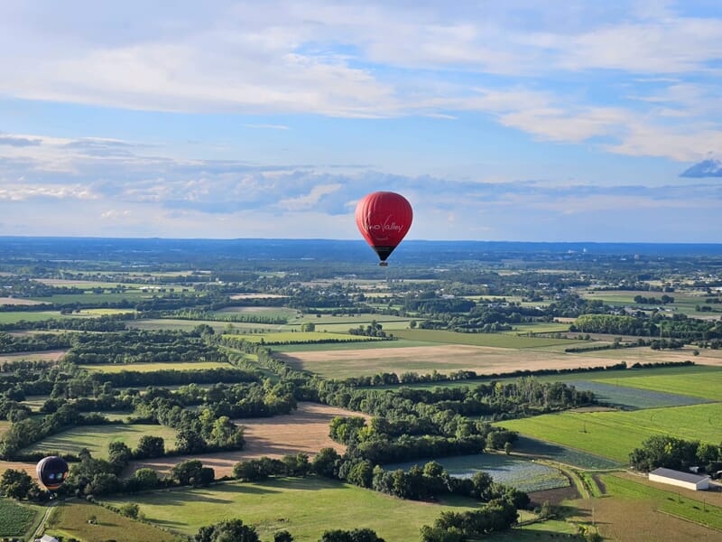 Montgolfière rouge volant au-dessus de champs et forêts dans un paysage rural étendu sous un ciel bleu.