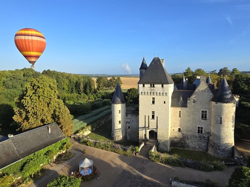 Montgolfière orange volant près d'un château médiéval entouré d'arbres verts sous un ciel bleu clair.