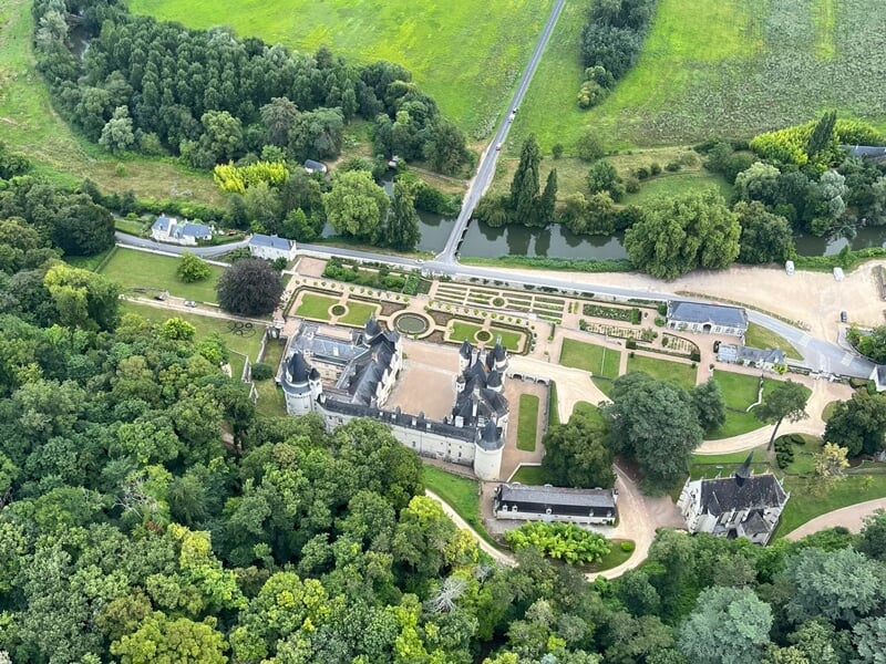 Vue aérienne d'un château avec jardins symétriques et allées, entouré d'une dense forêt verdoyante.