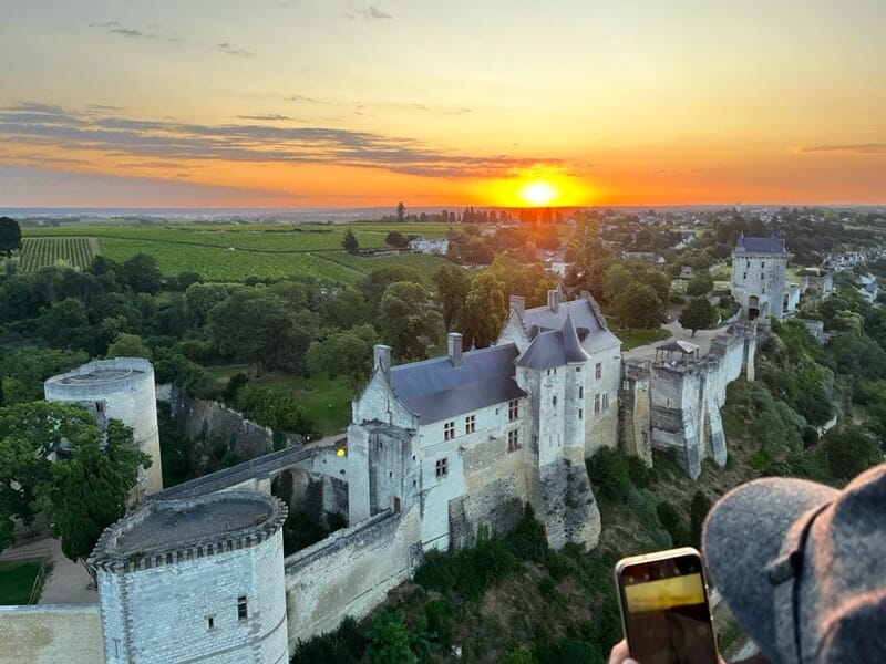 Coucher de soleil derrière un château fortifié avec des remparts, des vignes et une personne prenant une photo.
