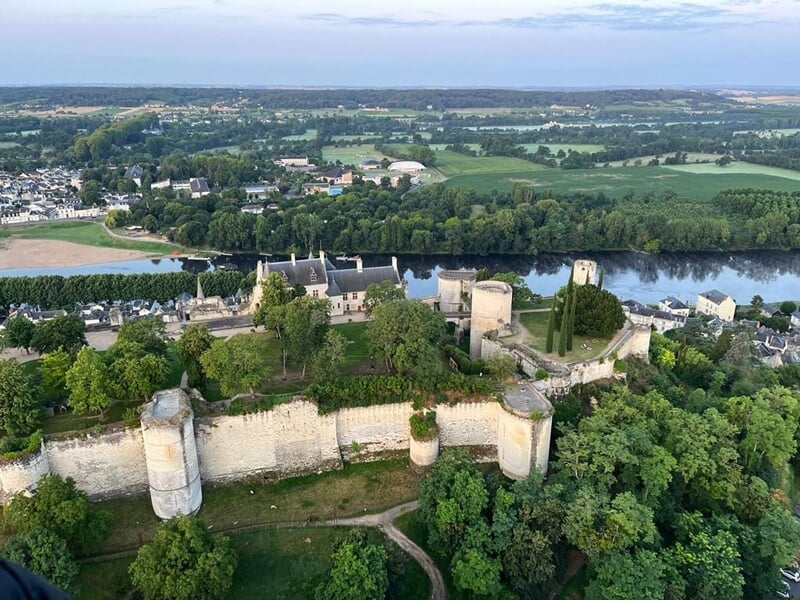 Vue aérienne d'un château fort entouré de murs et de tours, avec une rivière et une ville en arrière-plan.
