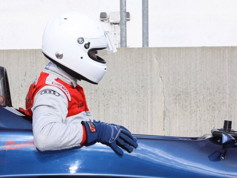 Pilote en combinaison rouge et casque blanc assis dans une monoplace bleue, prêt à prendre le départ.