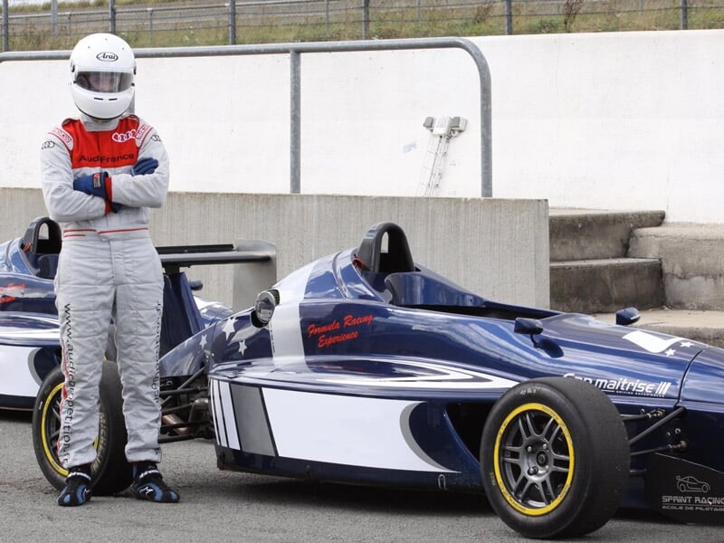Pilote en combinaison rouge et casque blanc debout à côté d'une monoplace bleue sur le paddock.