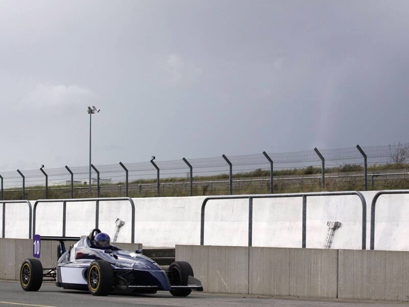 Monoplace bleue sur circuit, avec un pilote casqué en pleine accélération sur la ligne droite.