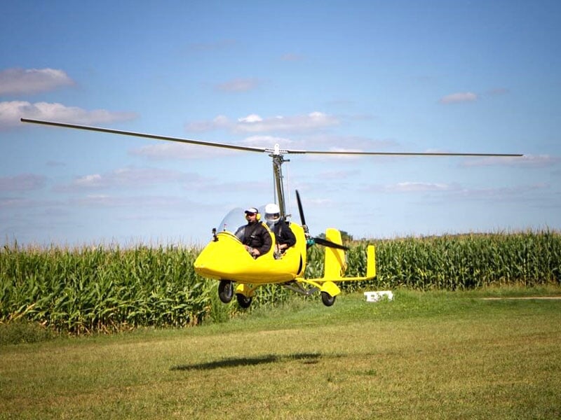 Autogire jaune atterrissant sur une pelouse verdoyante, avec des champs de maïs en arrière-plan et un ciel dégagé