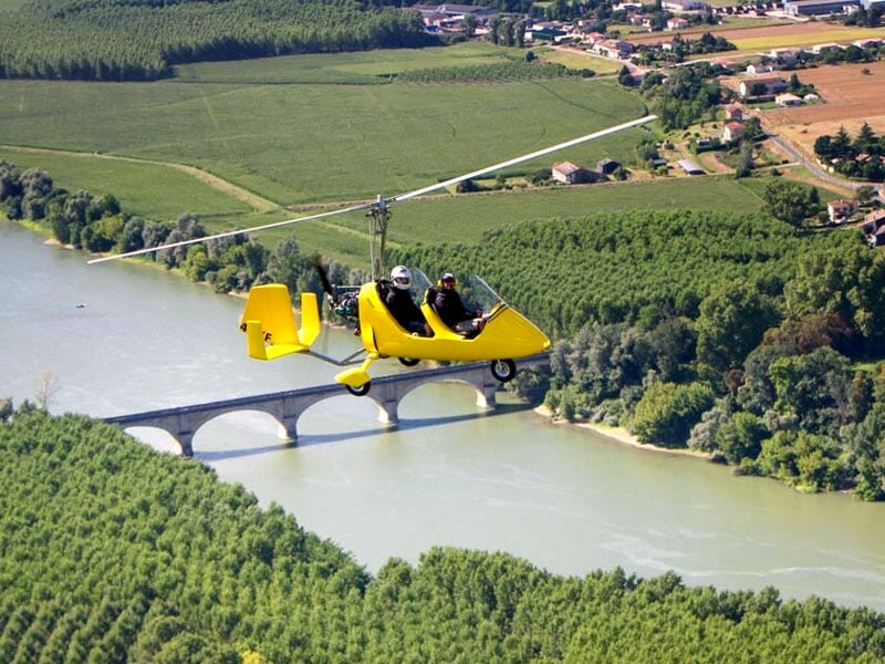 Autogire jaune survolant une rivière et un pont en pierre, entouré de champs verts et de forêts