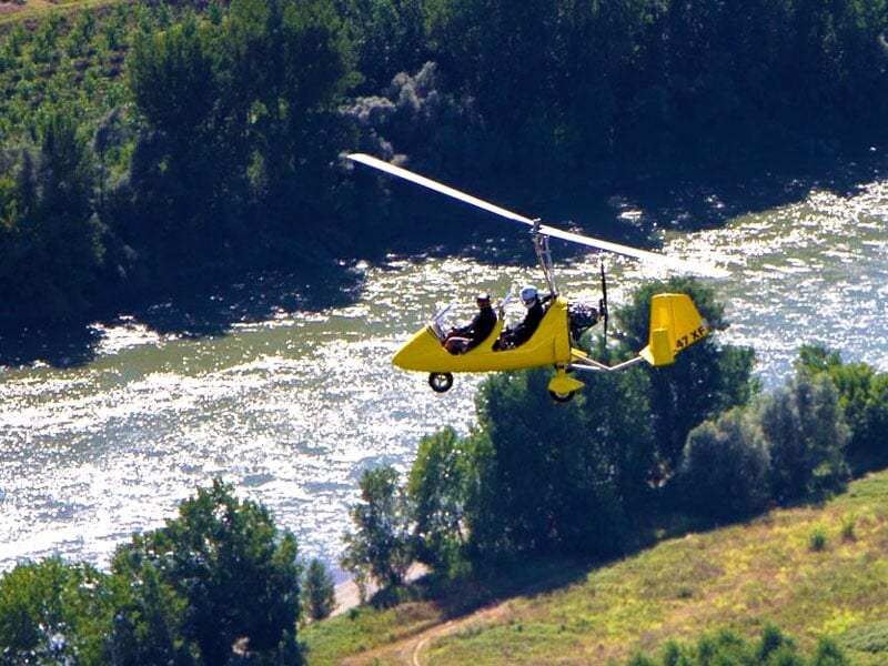 Autogire jaune évoluant à basse altitude au-dessus d’une rivière sinueuse entourée de végétation dense
