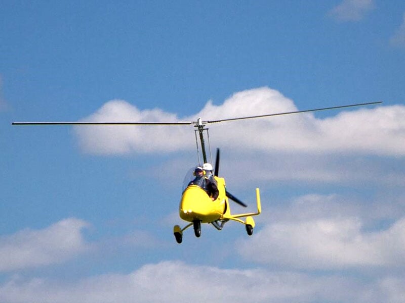 Autogire jaune en plein ciel, volant face à un ciel bleu parsemé de nuages blancs