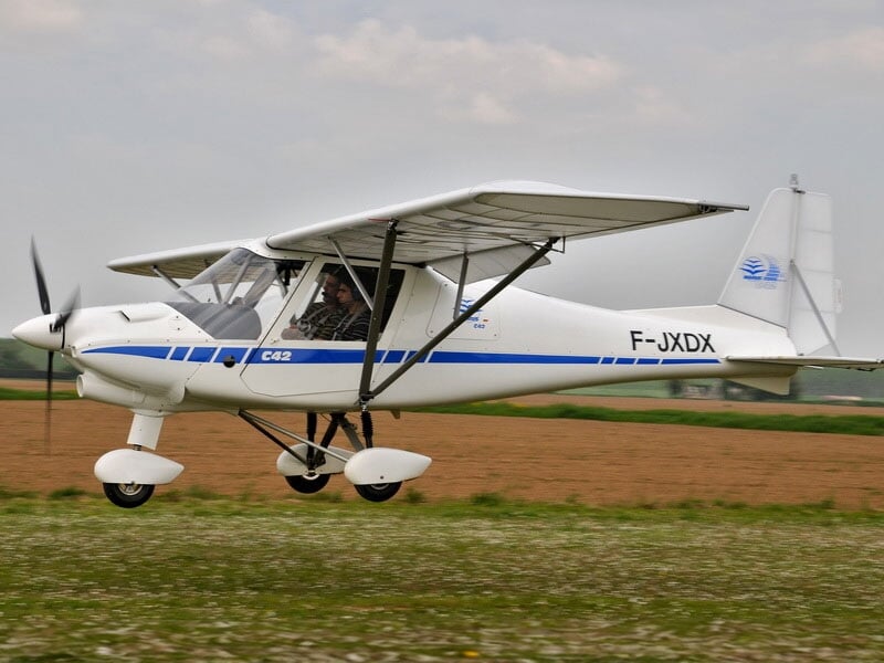 Avion ultraléger blanc C42 immatriculé F-JXDX en déplacement sur un terrain herbeux, hélice en rotation et arrière-plan flou de champs agricoles