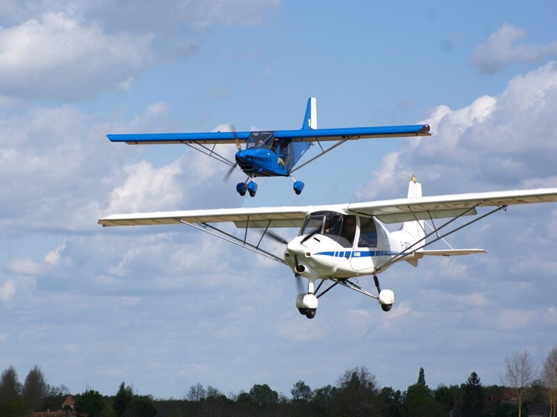 Deux petits avions légers en vol dans un ciel bleu avec quelques nuages blancs.
