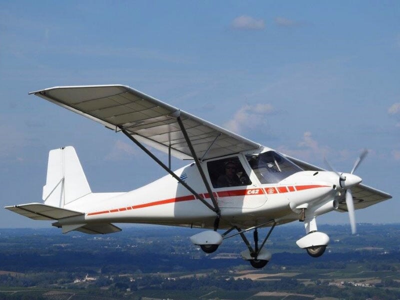 Avion ultraléger blanc modèle C42 en vol, ciel bleu dégagé avec quelques nuages et paysage rural en arrière-plan