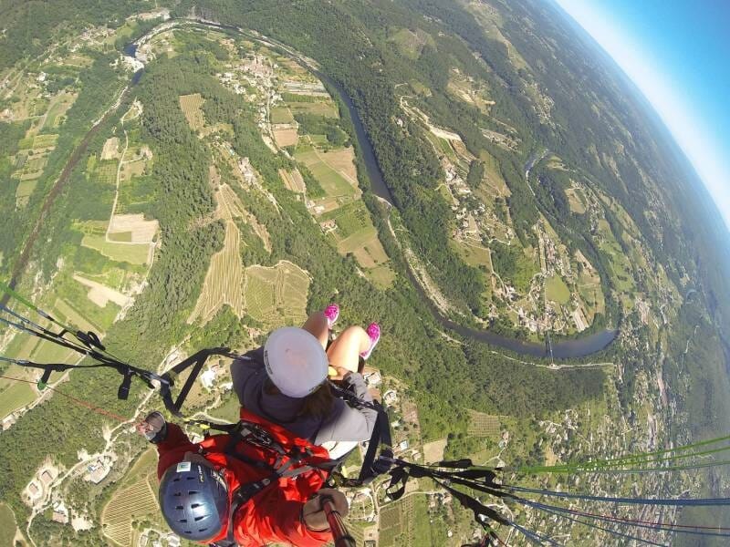 Parapente en tandem survolant un paysage vallonné avec forêts, vignobles et méandres d'une rivière sous un ciel dégagé.