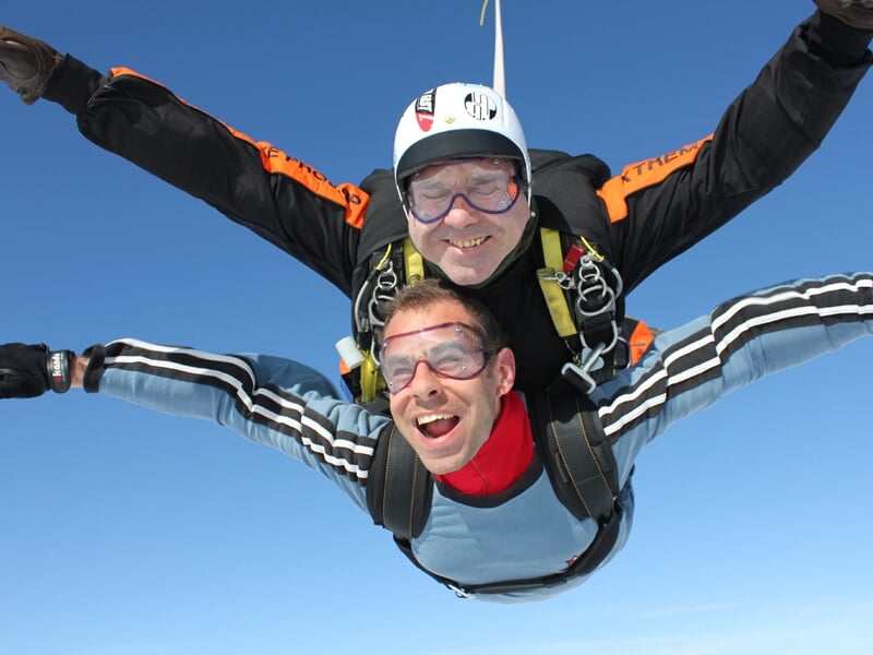 Saut en parachute en tandem, homme souriant en combinaison bleue et jaune, ciel bleu clair en arrière-plan.