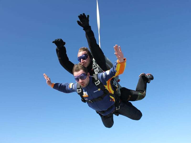 Deux parachutistes en tandem en chute libre, bras levés, dans un ciel bleu clair sans nuages.