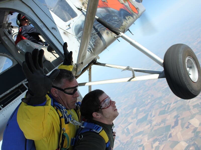 Parachutiste en tandem prêt à sauter d'un avion, ciel bleu et paysage agricole visible en dessous.