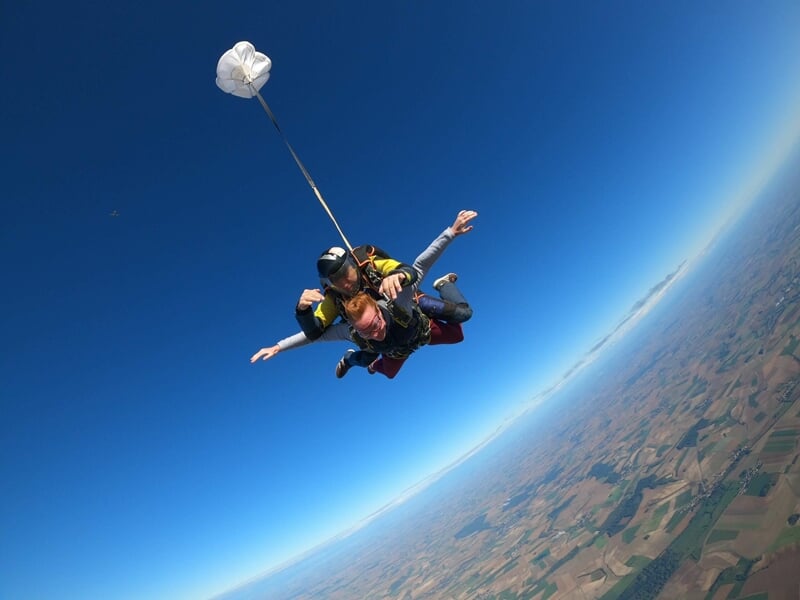 Saut en parachute en tandem avec parachute déployé, vue sur la campagne et ciel bleu clair.