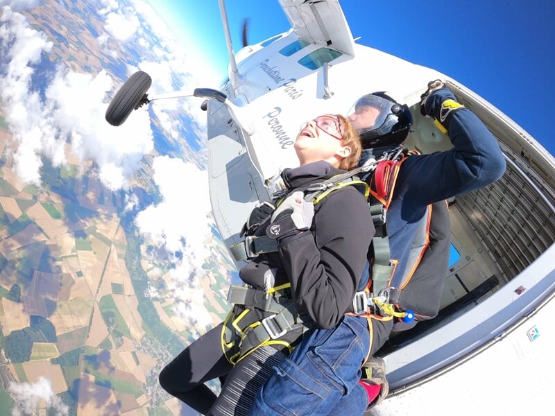 Femme souriante prête à sauter en parachute en tandem, attachée à un instructeur, ciel bleu et nuages.