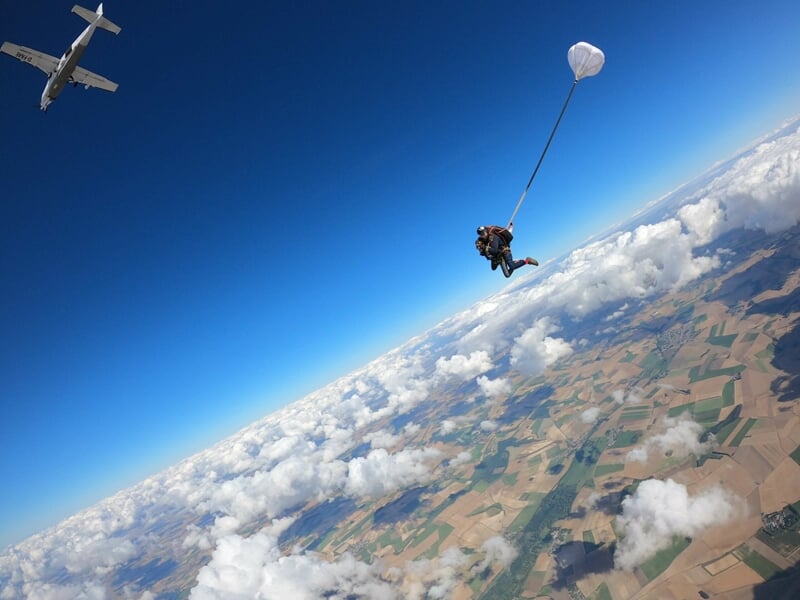 Deux parachutistes en chute libre, un avion s'éloigne en arrière-plan, ciel bleu et nuages blancs.