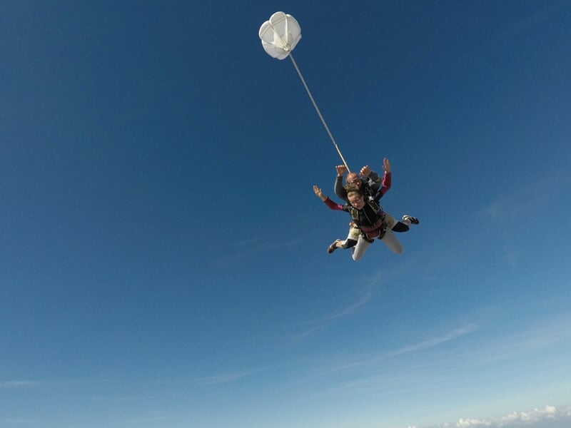 Parachutiste en tandem en chute libre, ciel bleu clair avec quelques nuages, paysage agricole en dessous.