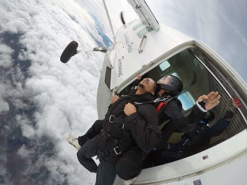 Deux personnes en combinaison de saut en parachute s'apprêtent à sauter d'un avion au-dessus des nuages, vue en plongée.