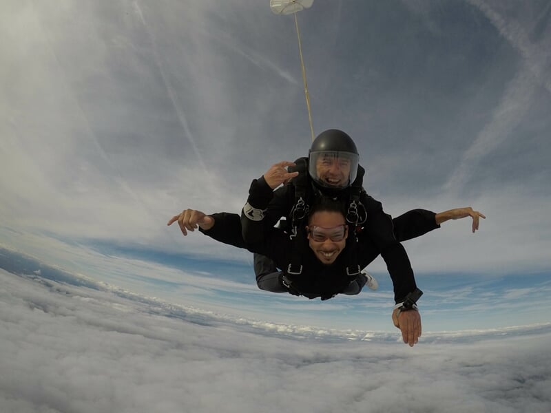 Deux parachutistes en tandem en chute libre, bras levés, ciel bleu clair et nuages blancs en arrière-plan.