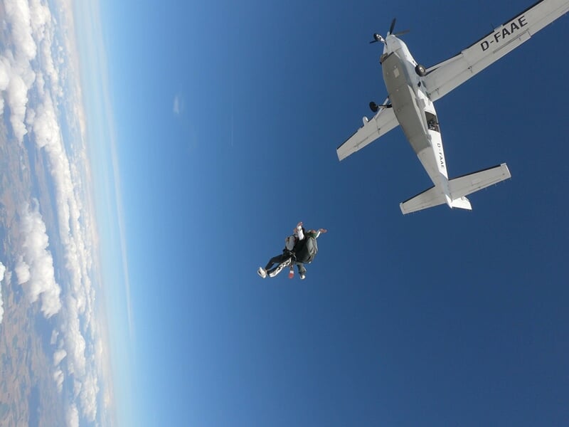 Saut en parachute en tandem, deux personnes en chute libre au-dessus d'un paysage rural sous un ciel bleu.