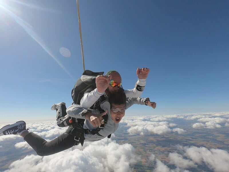 Parachutistes en tandem en chute libre, ciel bleu clair, paysage agricole visible en dessous, sensation de vitesse.