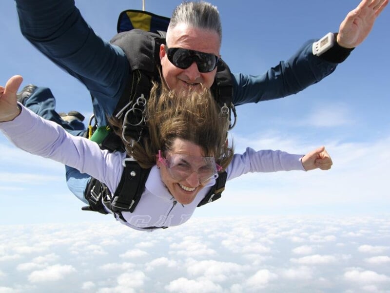 Parachutiste en tandem en chute libre, ciel bleu clair, paysage rural en dessous, sensation de liberté.