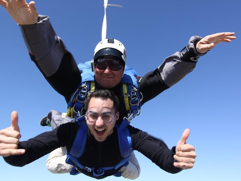 Saut en parachute en tandem, deux personnes en chute libre au-dessus d'un paysage rural sous un ciel bleu.