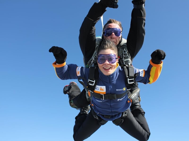 Deux personnes en tandem effectuent un saut en parachute sous un ciel bleu clair, souriantes et équipées de lunettes.