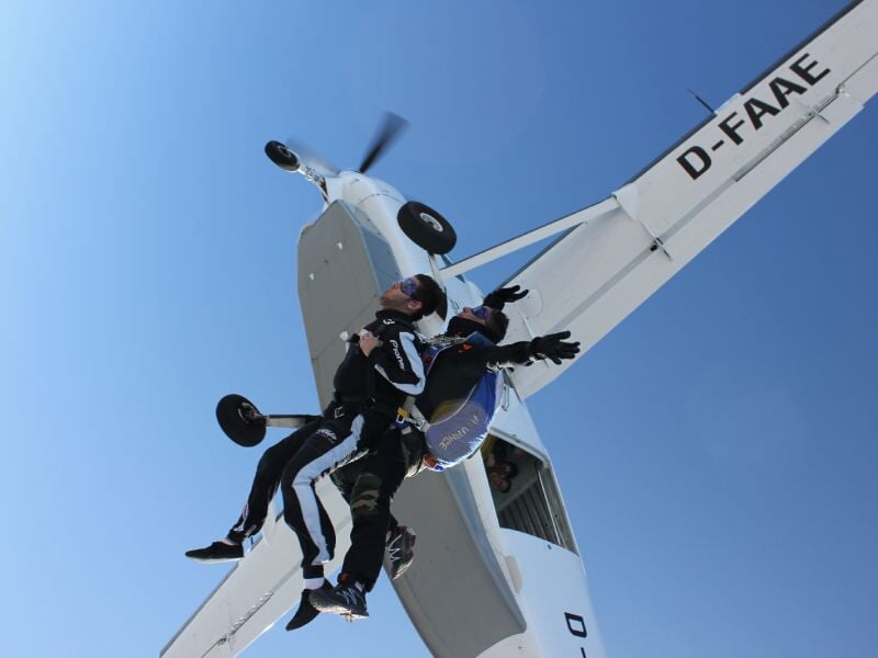 Deux parachutistes en combinaison noire sautant d'un avion blanc en plein ciel dégagé, en chute libre.