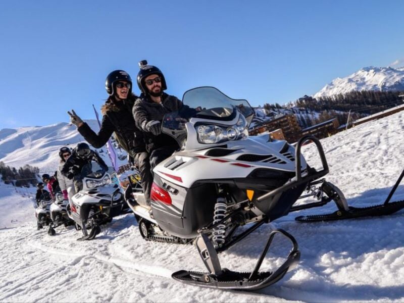 Deux personnes souriantes en tenue de neige conduisent une motoneige blanche sur un sentier enneigé en montagne.