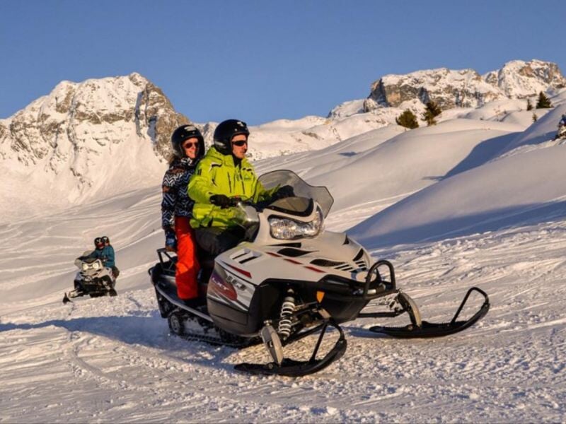 Deux personnes en vêtements d'hiver colorés conduisent une motoneige blanche sur une piste enneigée avec montagnes en arrière-plan.
