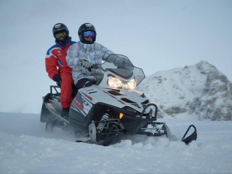 Deux conducteurs en combinaison de neige sur une motoneige blanche avancent sur une piste enneigée avec montagnes en arrière-plan.
