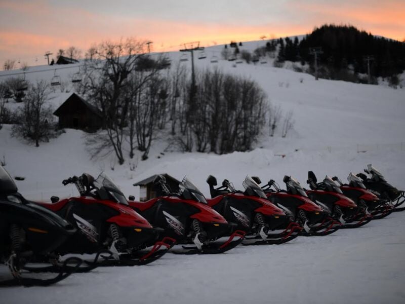 Plusieurs motoneiges rouges et noires alignées sur la neige au crépuscule avec des arbres et une colline.