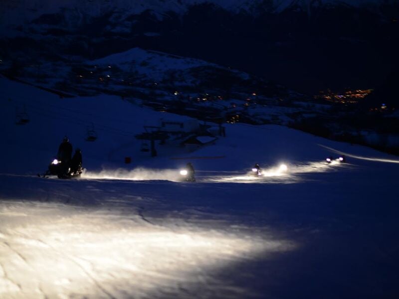 Motoneiges en mouvement la nuit sur la neige avec leurs phares allumés et un paysage montagneux en arrière-plan.