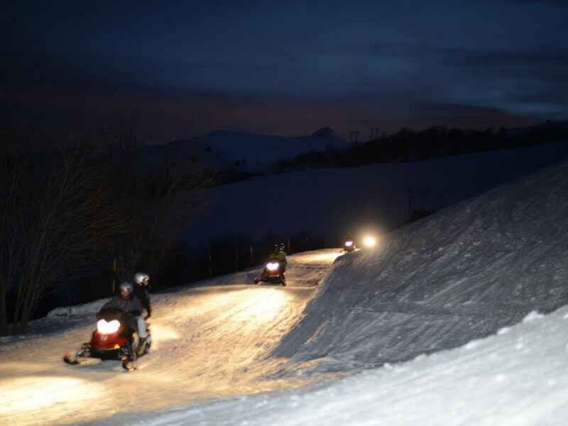 Deux personnes conduisent des motoneiges la nuit sur une piste enneigée éclairée par les phares dans un paysage vallonné.