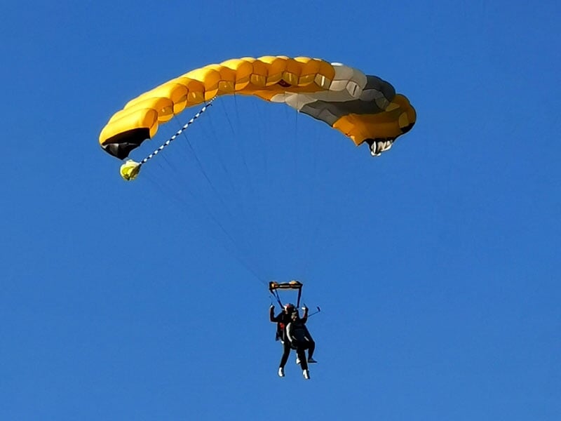 Un parachutiste descend avec un parachute jaune et noir ouvert, ciel bleu clair sans nuages.