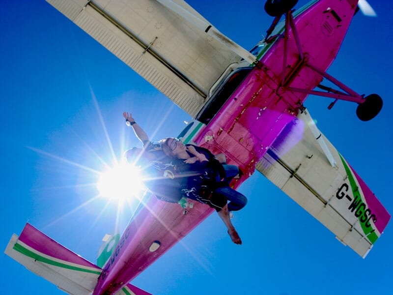 Une femme en parachute saute d'un avion rose avec le soleil éclatant derrière elle dans un ciel bleu.