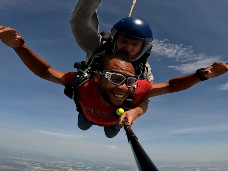 Deux personnes en parachute en tandem, souriantes, volant dans le ciel bleu au-dessus d'un paysage dégagé.