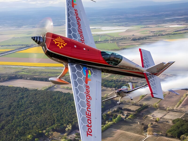 Vue en contre-plongée d'un avion acrobatique réalisant un virage serré avec un autre avion en arrière-plan, montrant les logos des sponsors sur les ailes