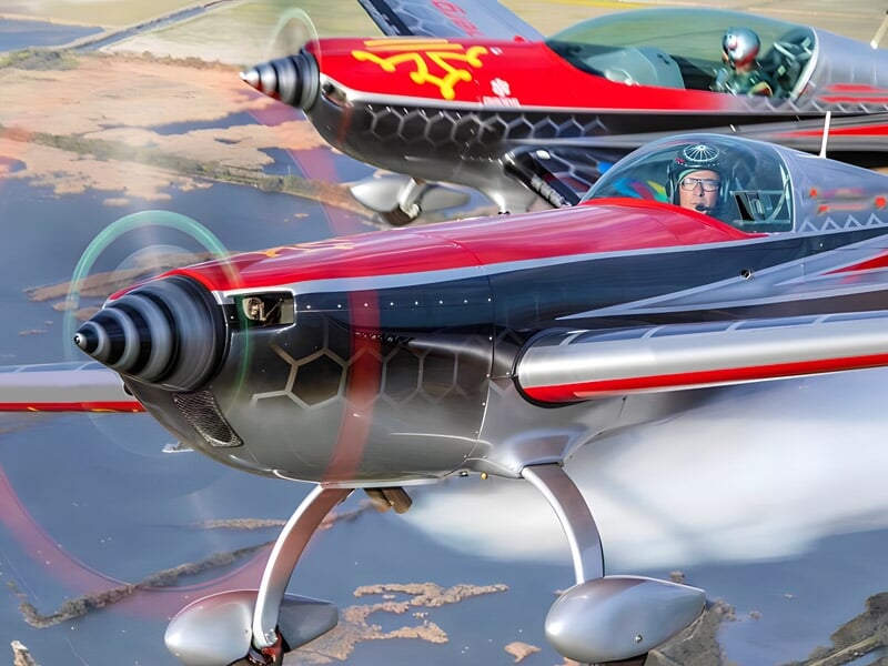 Vue rapprochée d'un avion acrobatique avec un pilote visible dans le cockpit, volant à basse altitude au-dessus d'un paysage d'eau et de terres